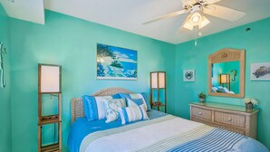 A bedroom with a bed, a ceiling fan, a painting, a mirror, and a dresser.