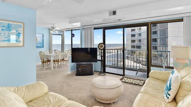 A spacious living room with a large flat-screen TV, a dining area with chairs, a round ottoman, and a balcony with a view of the sea.