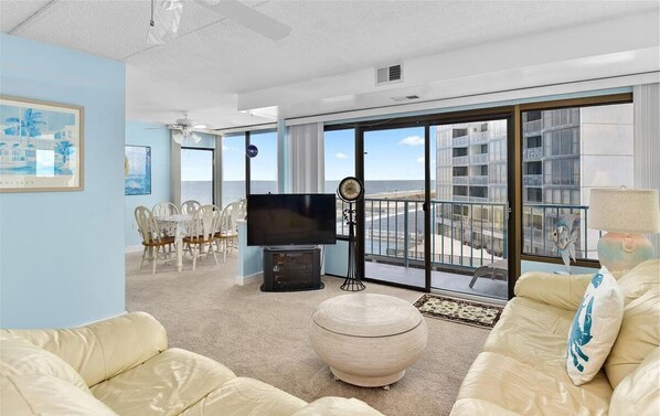 A spacious living room with a large flat-screen TV, a dining area with chairs, a round ottoman, and a balcony with a view of the sea. - XPLORIE ACTIVITIES & LINENS INCLUDED*!  2BR/2BA Capri condo with ocean & bay views! (Ocean City)