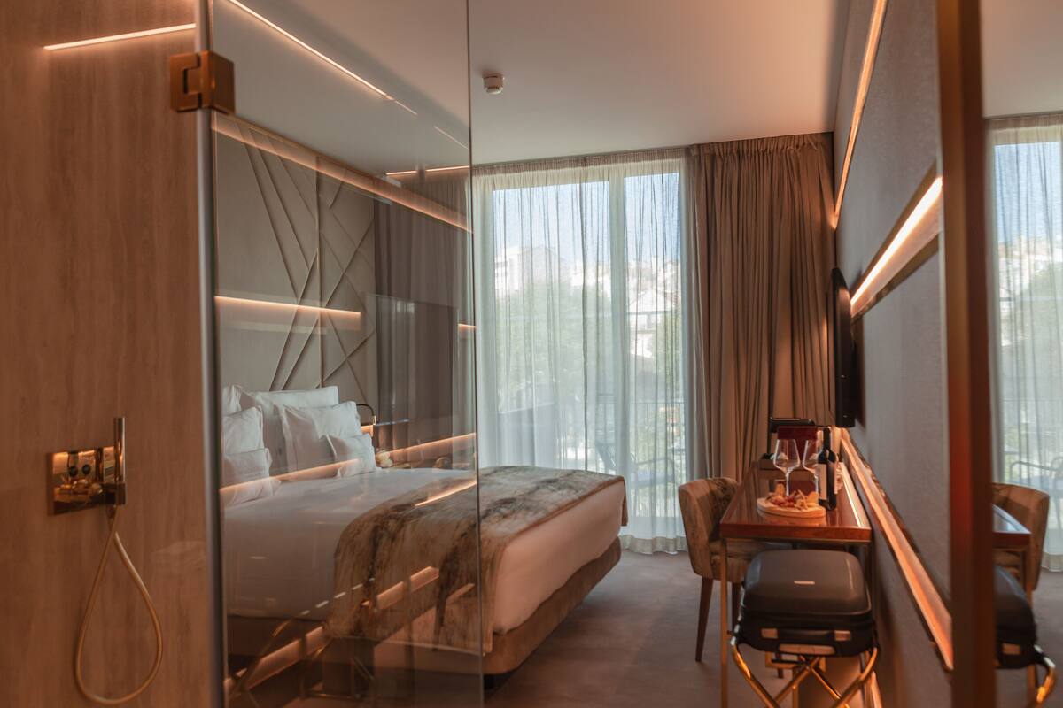 Une chambre d’hôtel moderne dotée d’un lit entouré de parois de verre, d’un bureau avec une chaise et offrant une vue sur la ville grâce à de grandes fenêtres.