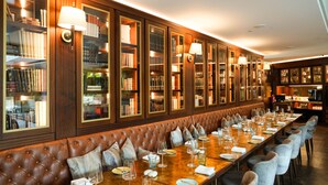 Una larga mesa de comedor con sillas, un banco de cuero y estanterías con lámparas.