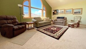 A spacious living room with a high ceiling, large windows, and a variety of seating options including a brown sofa, a beige sofa, and a white armchair. - Windsor Drive Monthly Rental (Cody)