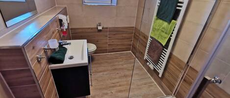 Une petite salle de bain avec un plancher en bois, un lavabo blanc, des toilettes, un espace douche et une fenĂŞtre.