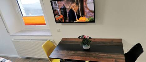 Un coin repas avec une table en bois, des chaises jaunes et noires, une plante en pot et un téléviseur fixé au mur qui diffuse une scène.