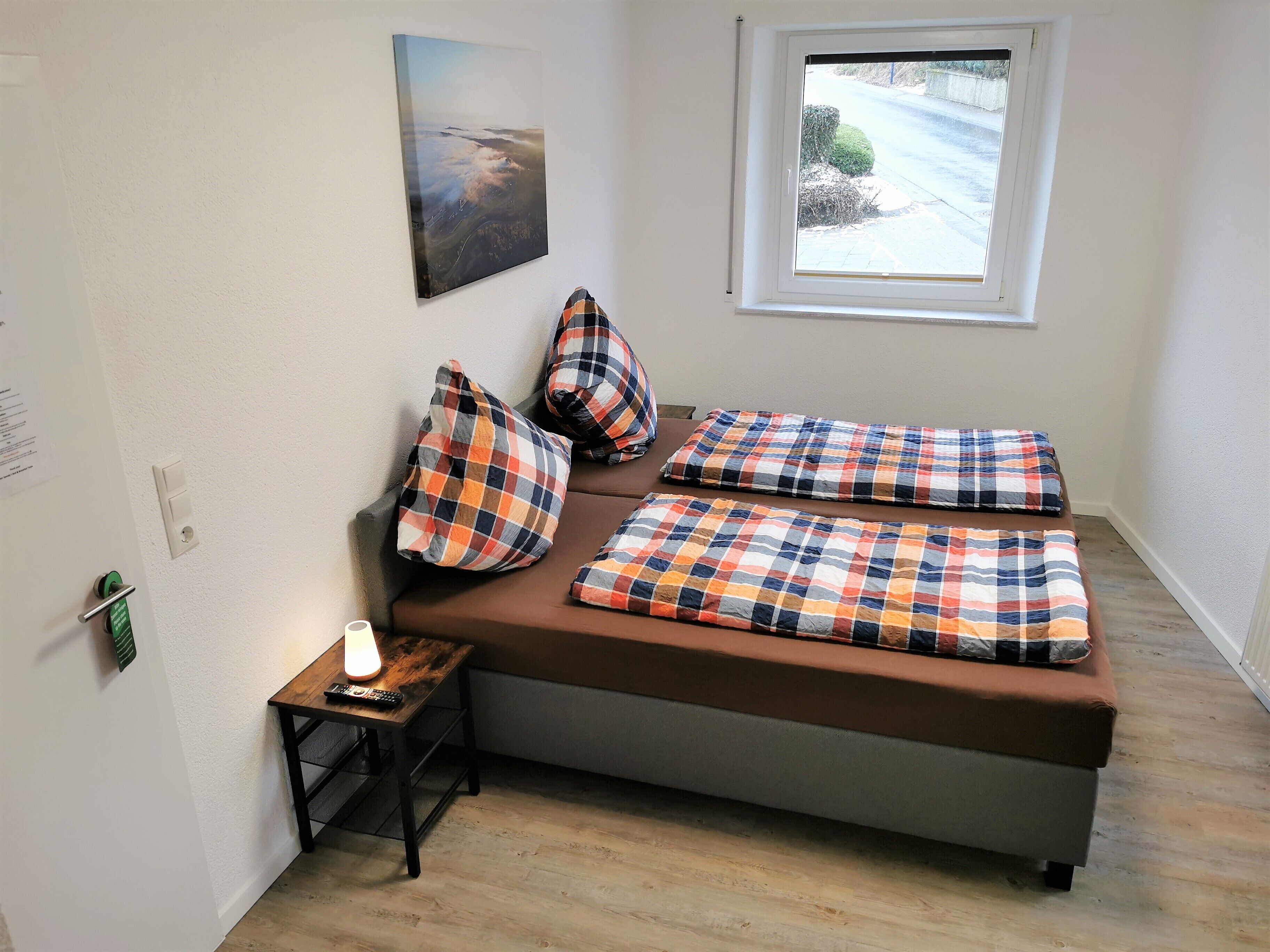 Una habitación pequeña y acogedora con una cama, una ventana, una lámpara y una mesita con un control remoto.