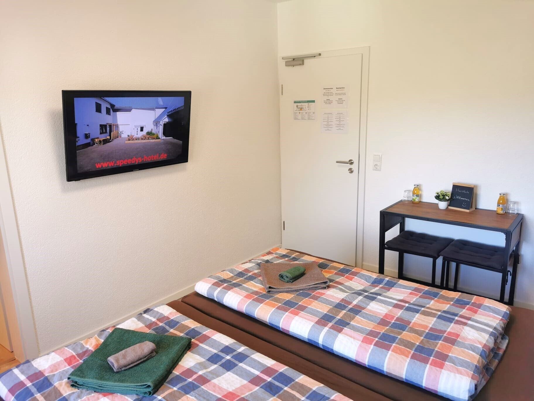 Una habitación con una cama, una mesita con un cartel y un televisor que muestra la página web de un hotel.