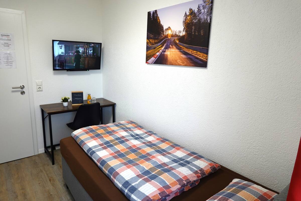 A small, neatly arranged room with a bed, a desk, a chair, a television, and a framed picture on the wall.