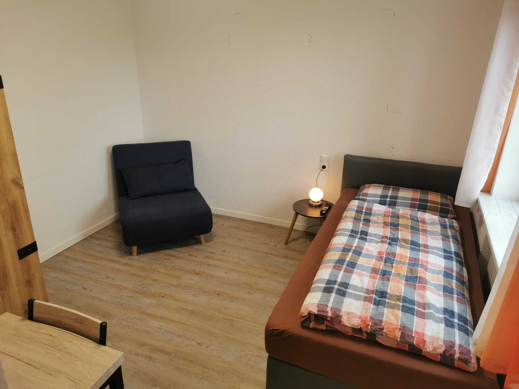 Una habitación pequeña con una cama, una silla, una mesita de noche con una lámpara y una ventana.