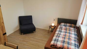 Una habitación pequeña con una cama, una silla, una mesita de noche con una lámpara y una ventana.