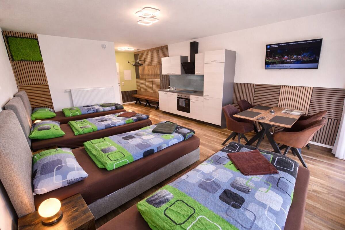 A modern hotel room with a sofa bed, a dining table with chairs, a TV, and a kitchen area.