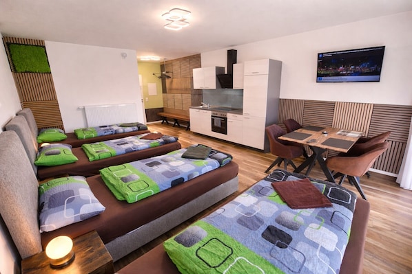 A modern hotel room with a sofa bed, a dining table with chairs, a TV, and a kitchen area.