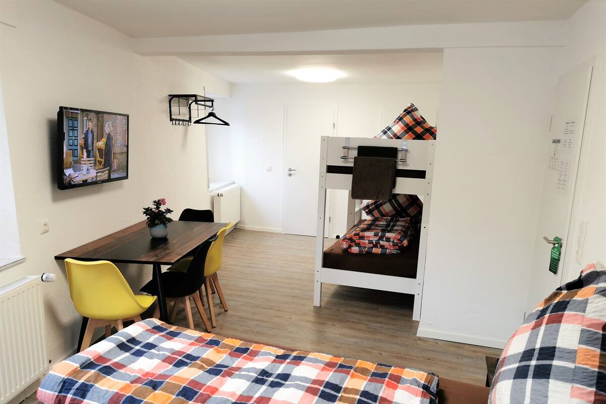 A dormitory room with a bunk bed, a dining table with chairs, a TV mounted on the wall, and a bed with a colorful blanket.