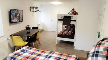 Une chambre de dortoir avec un lit superposé, une table à manger avec des chaises, un téléviseur fixé au mur et un lit recouvert d’une couverture colorée.