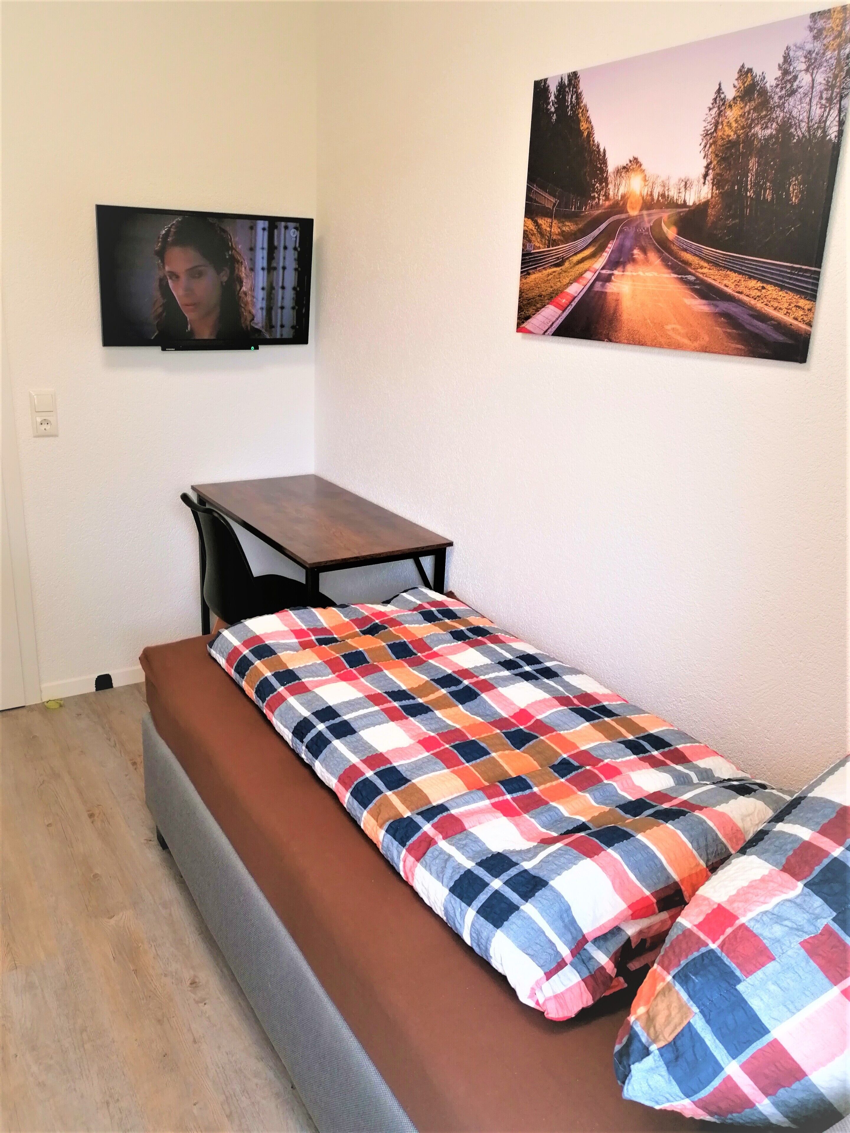 A small, single-bed room with a wooden table and bench, a colorful checkered bedspread, and two wall-mounted pictures.