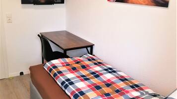 A small, single-bed room with a wooden table and bench, a colorful checkered bedspread, and two wall-mounted pictures.