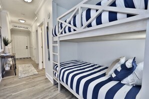Uma cama de beliche com roupa de cama listrada em azul marinho e branco, uma escada de madeira e uma pequena mesa com uma planta. Uma placa na parede diz "OUR HAPPY PLACE".