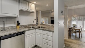 A kitchen with white cabinets, granite countertops, a dishwasher, and a dining area with a table and chairs. - Sunset Rentals | St. Andrews 1662 (Hilton Head Island)