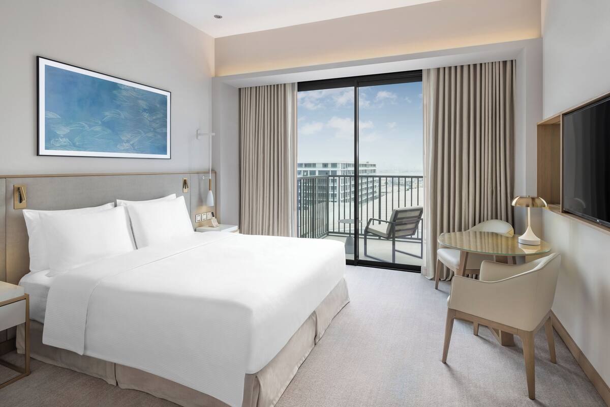 Quarto de hotel com uma cama grande, uma escrivaninha com cadeira, televisão e varanda com vista para edifícios.