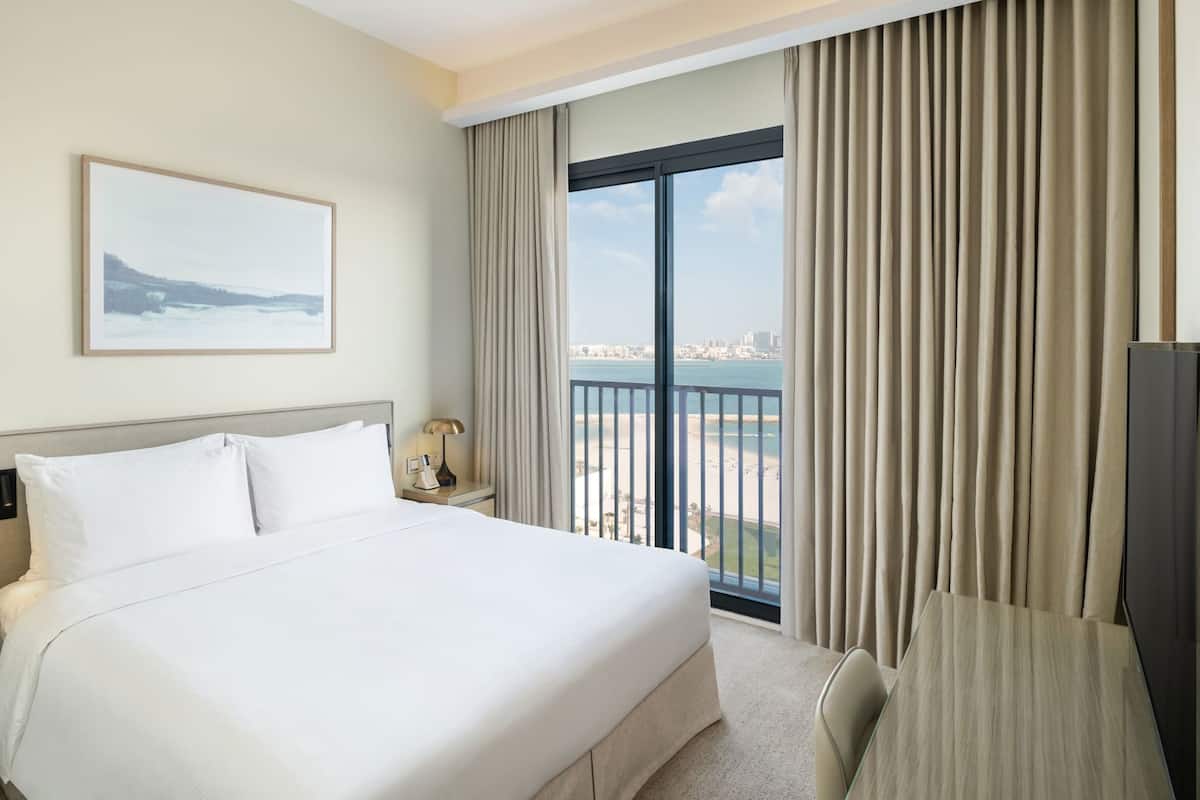 A hotel room with a bed, a desk, a chair, a nightstand, a lamp, a painting, and a view of the beach and city.