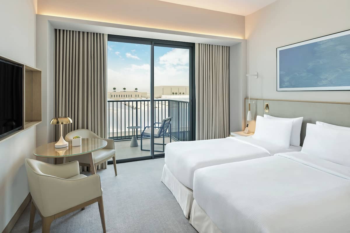 Quarto de hotel com duas camas, uma escrivaninha com cadeira, televisão e varanda com vista para edifícios.