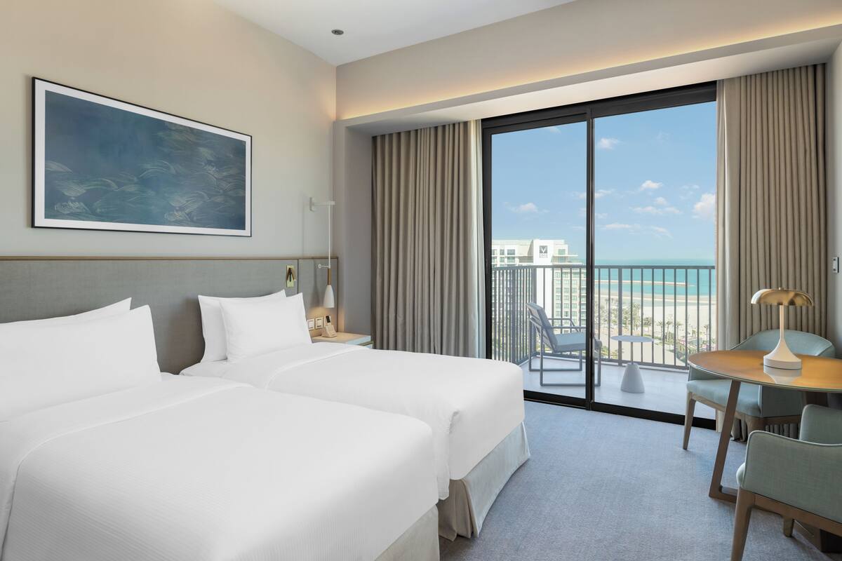 Quarto de hotel com duas camas, varanda com vista para a praia, área de refeições e um quadro na parede.