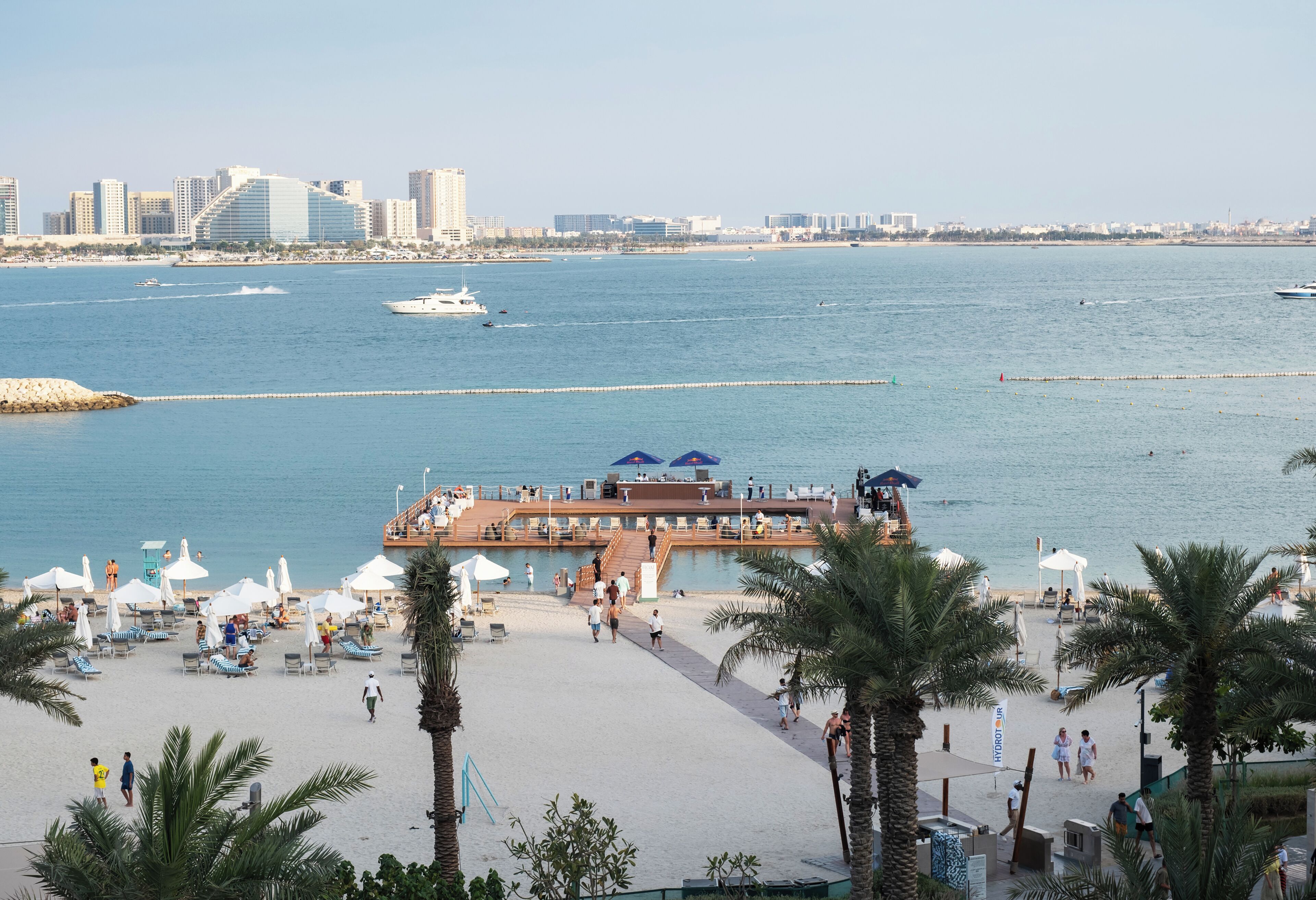 A beachside resort with a pool area, umbrellas, and a view of the sea and city skyline.