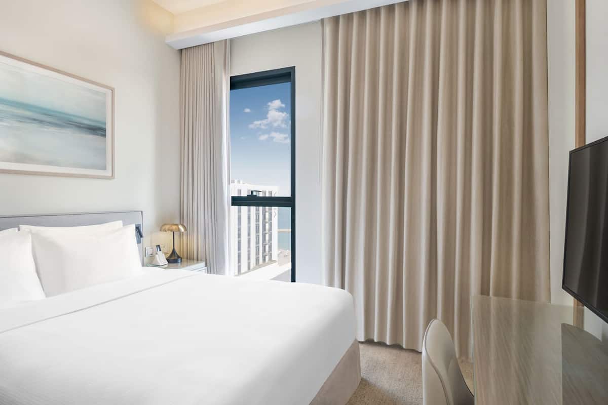 Quarto de hotel com uma cama grande, uma escrivaninha, televisão, janela com cortinas e um quadro na parede.