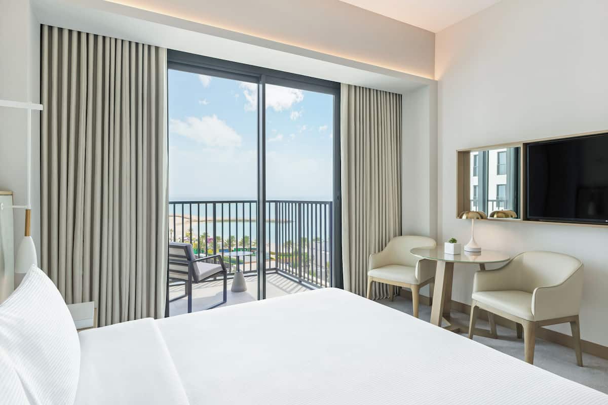 Quarto de hotel com uma cama grande, uma televisão, uma pequena mesa com duas cadeiras e uma varanda com vista para a cidade.