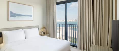 A hotel room with a large bed, a desk, a chair, a nightstand, a lamp, a framed picture on the wall, and a view of the city and sea through the window.