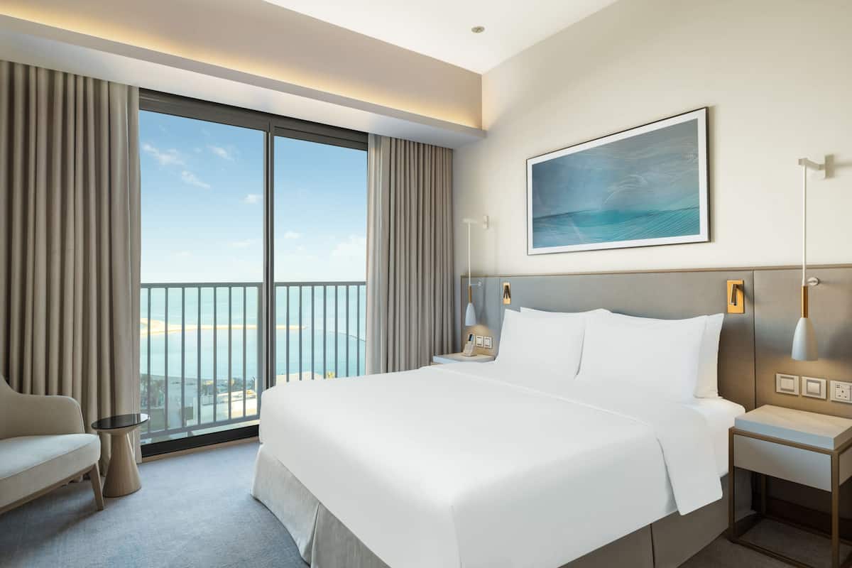 Quarto de hotel com cama grande, escrivaninha, cadeira, vista para o mar e um quadro na parede.