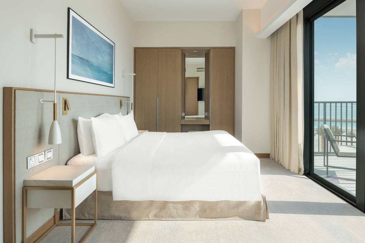 Quarto de hotel moderno com uma cama grande, uma mesa de cabeceira, uma escrivaninha, uma janela ampla com vista para uma varanda e um quadro na parede.