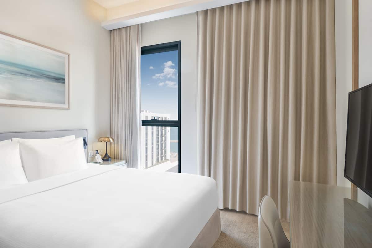 Quarto de hotel com uma cama grande, uma escrivaninha com televisão, uma mesa de cabeceira com abajur e uma janela com cortinas.