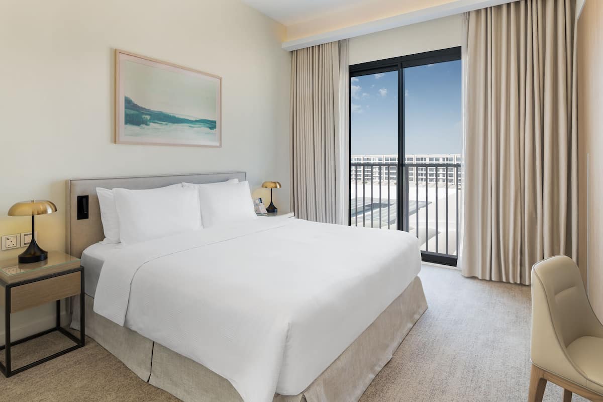 Quarto de hotel com uma cama grande, mesa de cabeceira com abajur, uma cadeira, um quadro na parede e vista da cidade pela janela.