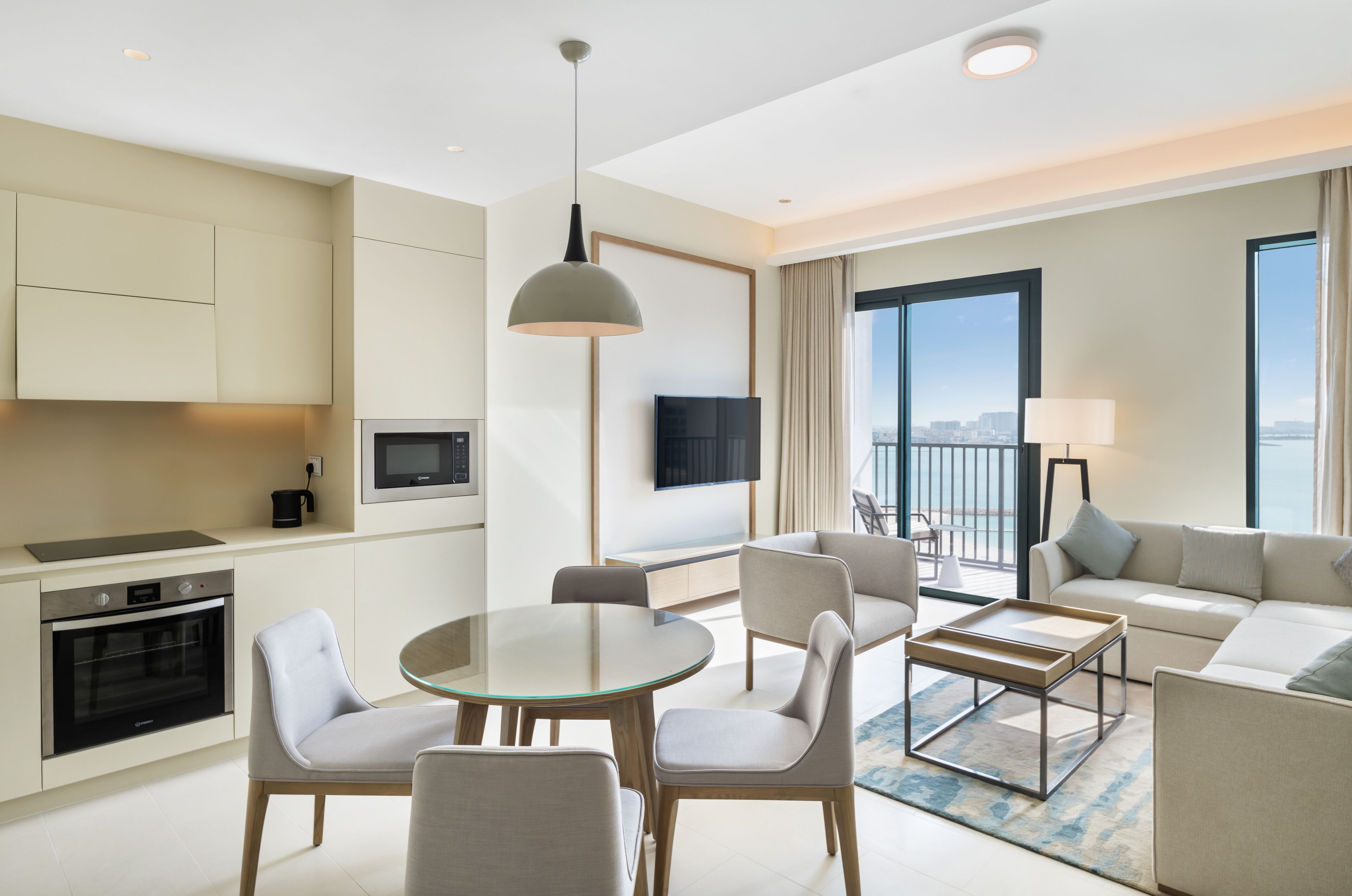 A modern hotel room with a kitchen, dining area, and living space. It includes a flat-screen TV, a sofa, and a balcony with a view.