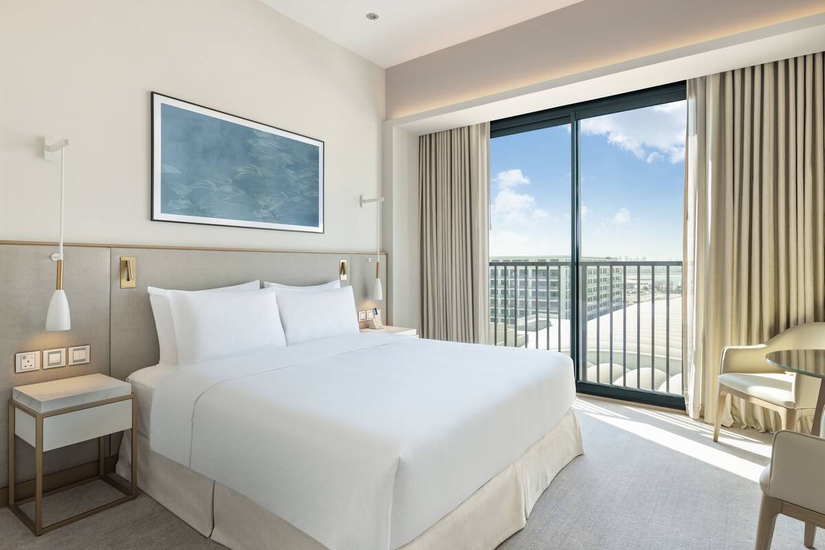 Quarto de hotel moderno com uma cama grande, uma mesa de cabeceira com abajur, uma obra de arte emoldurada na parede e vista para a cidade pela janela.