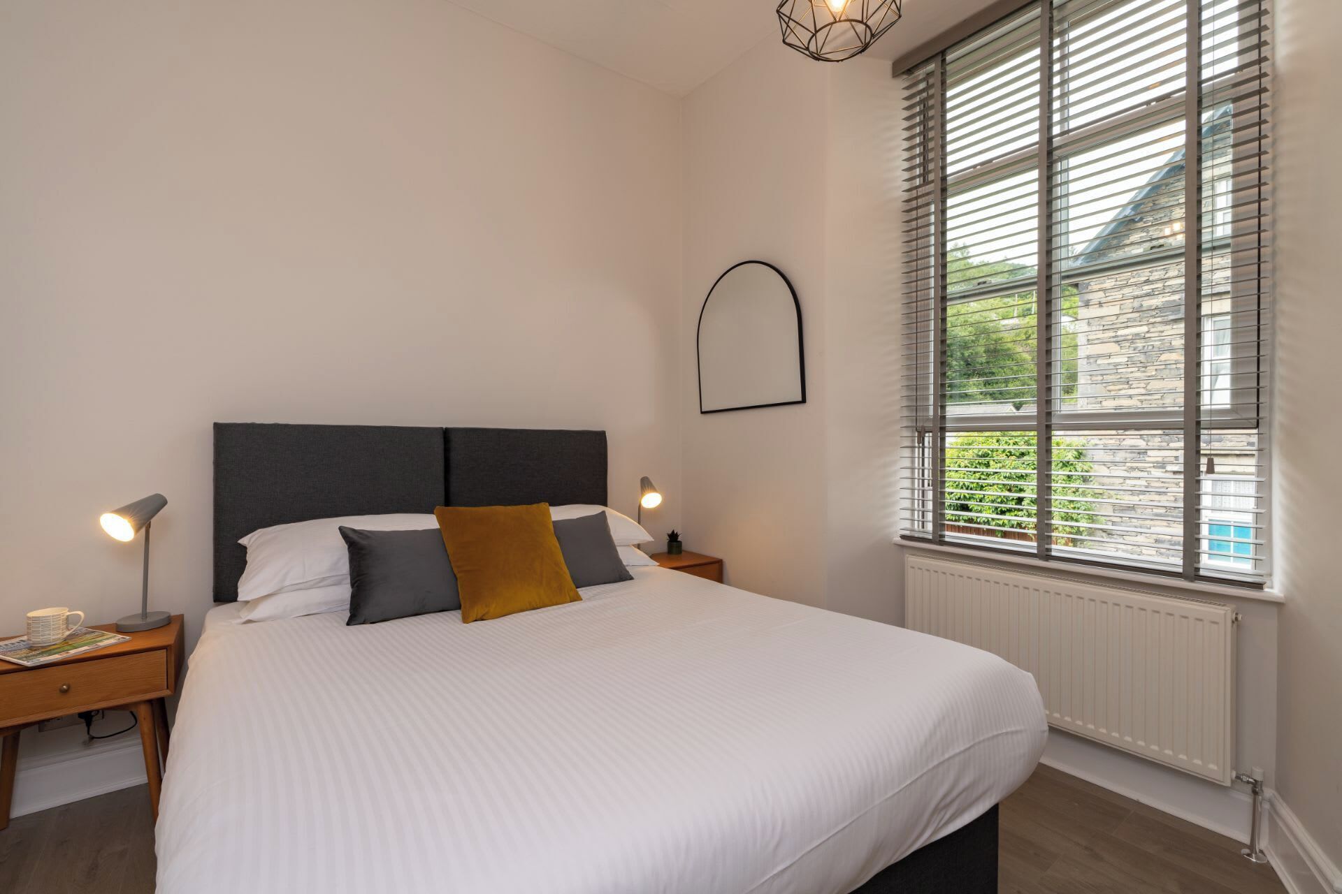 A neatly made bed with white bedding, a dark headboard, and two bedside lamps. A window with blinds is visible, offering a view of greenery outside.
