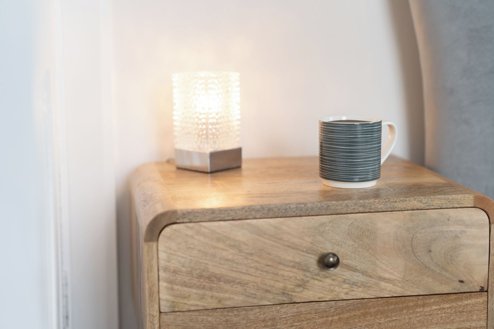 Una mesita de noche de madera con un cajón y una lámpara texturizada a su lado.
