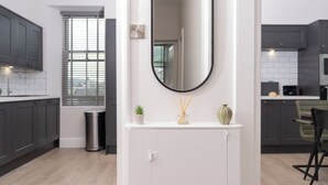 Une cuisine moderne avec des meubles de couleur sombre, un plan de travail blanc et un miroir placé au-dessus d’une petite étagère ornée d’une plante et d’un diffuseur de parfum à bâtonnets.