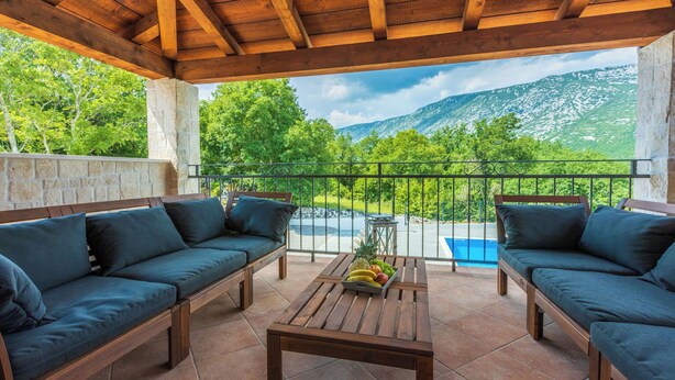 Ein überdachter Außenbereich mit Holztisch und zwei Sofas mit Blick auf eine Berglandschaft und einen Pool.