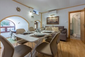 Een eetruimte met een witte tafel gedekt voor vier personen, omringd door beige stoelen. Er is een bank en een deur naar een douchegedeelte.