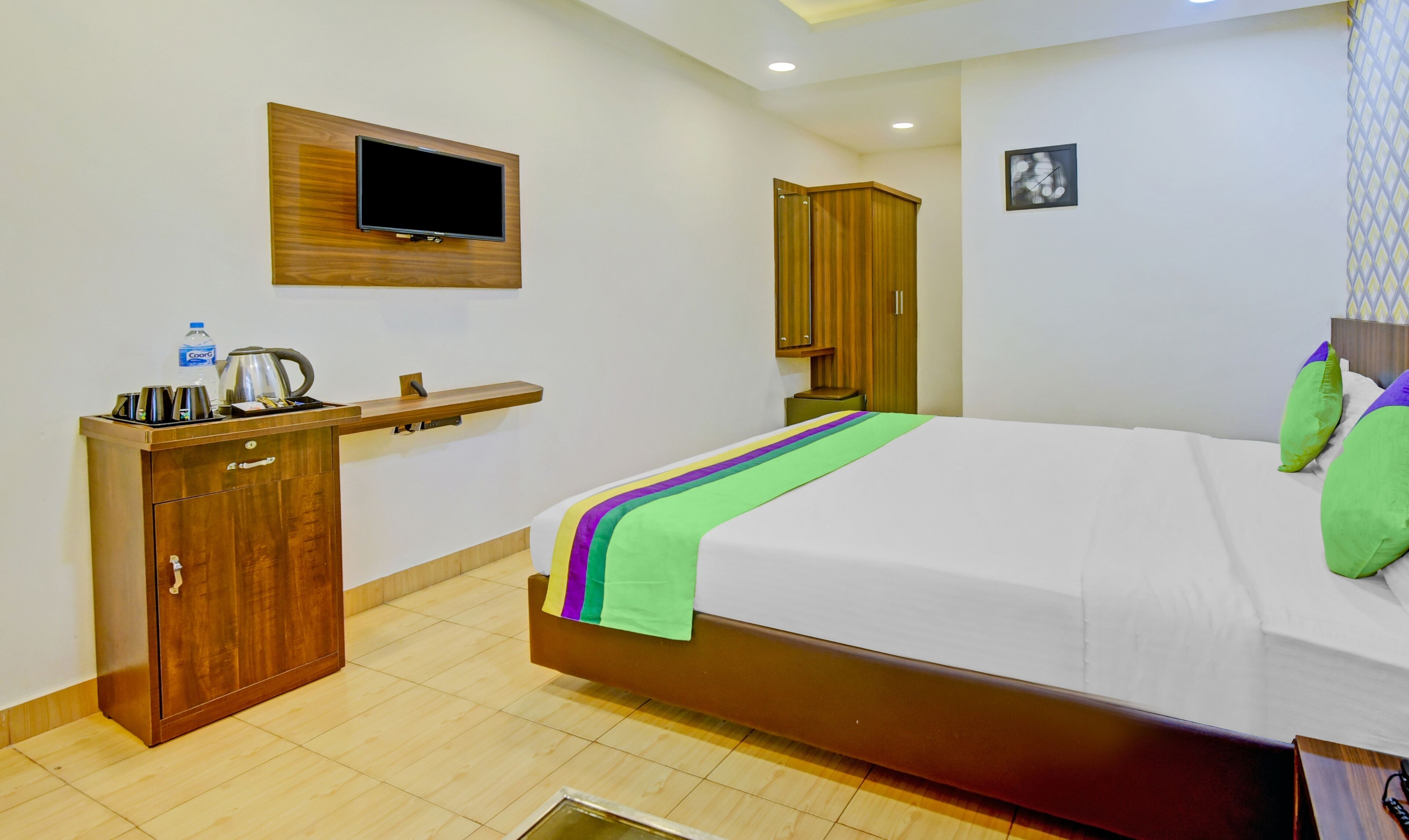 A hotel room with a bed, a wooden desk, a television mounted on the wall, and a wardrobe.