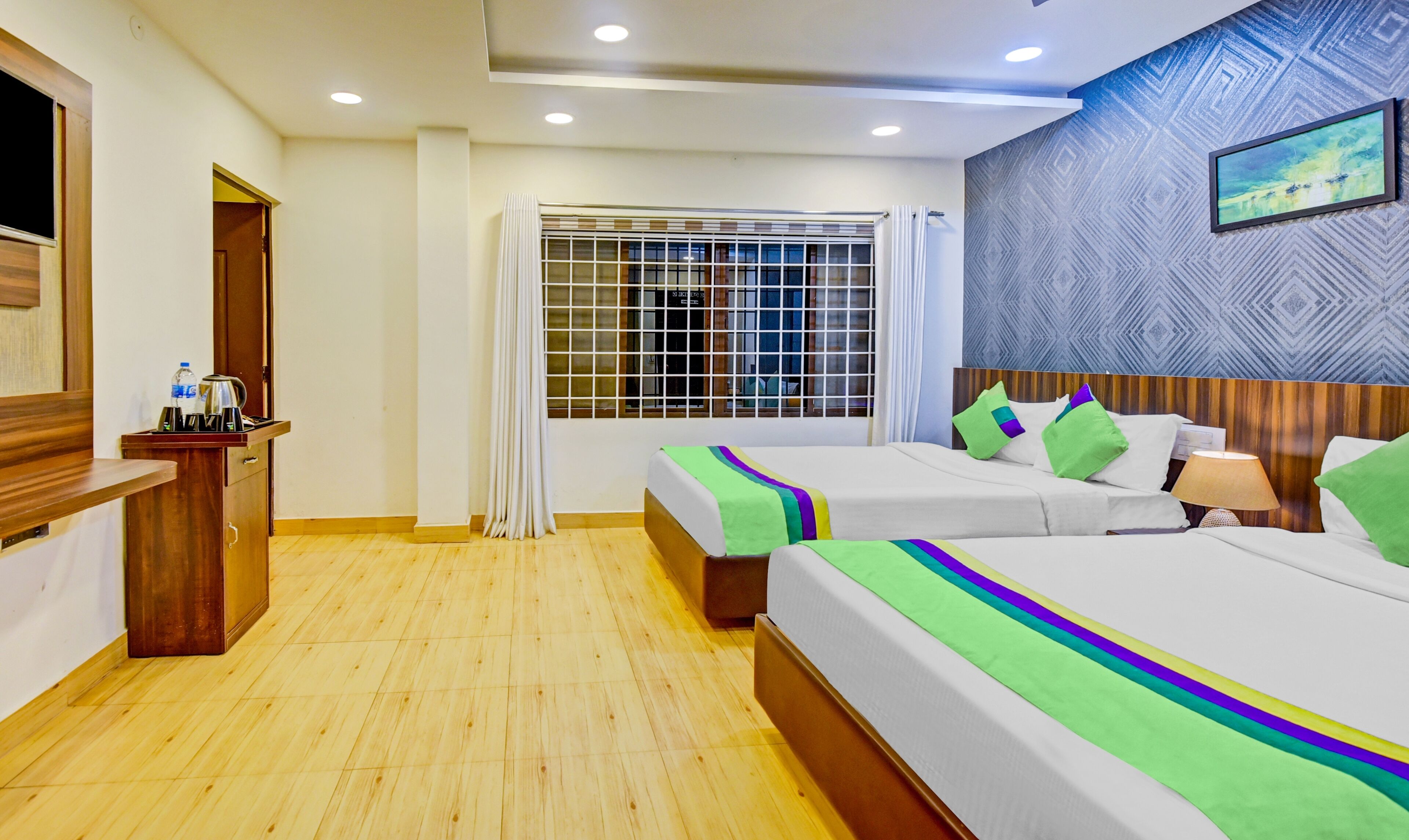 Habitación de hotel con dos camas, un escritorio de madera con hervidor y botella de agua, una ventana con cortinas y una pared con estampado.