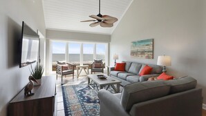 A modern living room with a large window overlooking the beach, a ceiling fan, and a cozy seating area with a gray sofa and armchairs.