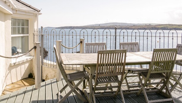 Une terrasse en bois avec une table et des chaises, donnant sur une étendue d’eau, et entourée d’une clôture en métal.