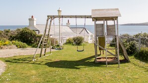 Un ensemble de jeux d’extérieur en bois, comprenant une petite structure en bois, une zone herbeuse et une vue sur la mer et les maisons au loin.