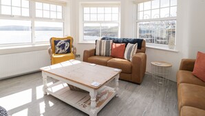 Un salon lumineux avec de grandes fenêtres, une table basse en bois, un fauteuil jaune et un canapé marron.