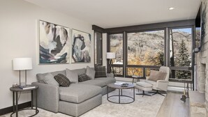 Ein modernes Wohnzimmer mit einem grauen Ecksofa, einem runden Couchtisch und einem beigen Sessel. Zwei große Fenster bieten Blick auf Bäume und ein Gebäude.