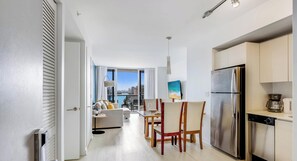A modern kitchen with a stainless steel refrigerator, a dining area with a table and chairs, and a living room with a sofa and a view of the city.