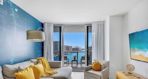 A modern living room with a sofa, armchair, and a view of a cityscape and water through large windows.