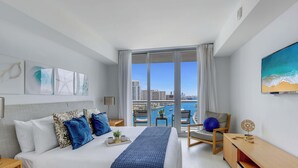 A modern hotel room with a large bed, a desk, a chair, and a view of a cityscape through a large window. - 2BR Apartment w/ Balcony - Near Beach (Hallandale Beach)